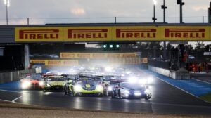 A line of race cars with their headlights on take a turn at a racetrack