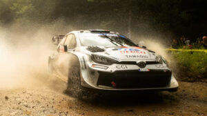 An FIA World Rally Championship rally car driving at high speed through a turn, kicking up dust