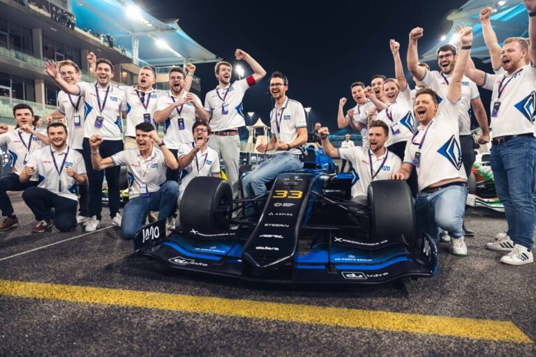 TUM team members celebrate their win at the second A2RL Grand Final with the car parked between them and the grandstand rising up on either side into the night sky