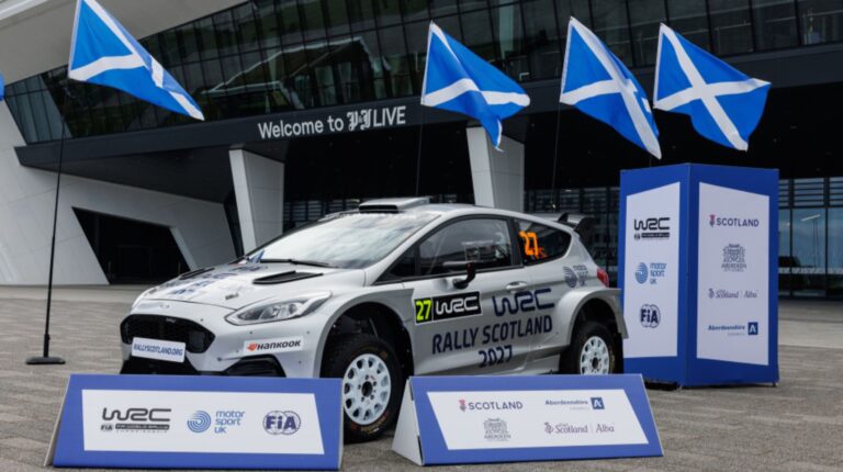 A WRC rally car in front of Scotland flags.